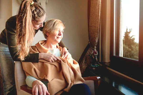 Eine junge Frau mit langem Zopf legt liebevoll eine Decke um die Schultern einer älteren Dame, die im Sessel neben einem Fenster sitzt. Sie teilen einen Moment der Fürsorge in einem warm beleuchteten Raum. Die ältere Frau, lächelnd und entspannt, blickt nach draußen, während die jüngere Frau sich zu ihr neigt und ein Gefühl von Geborgenheit und Zuneigung ausstrahlt. Diese Szene spiegelt die Fürsorglichkeit in der häuslichen Pflege wider.