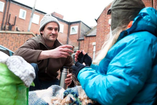 Ein junger Mann reicht einen Becher einer Thermoskanne an eine Person mit blauer Jacke